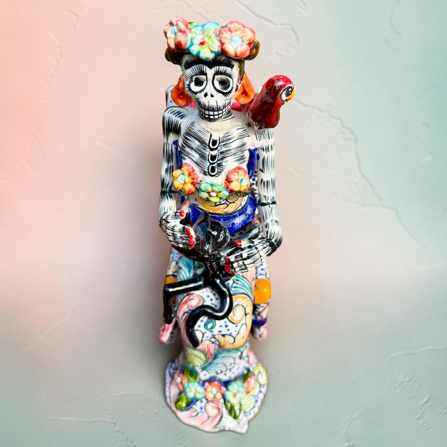 Frida Kahlo Talavera day of the dead figurine sitting in a chair holding a monkey with a parrot on her shoulder, top view looking down on chair.