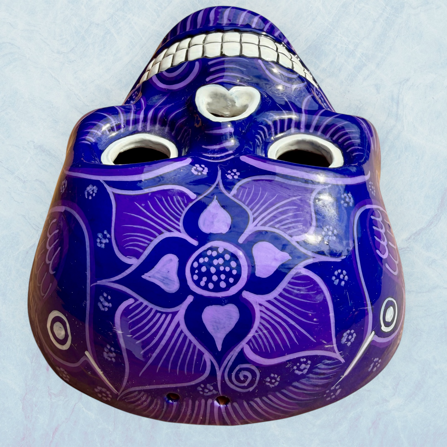 A clay, purple with designs, Mexican-style skeleton skull face mask hanging on a soft blue surface top forehead view.