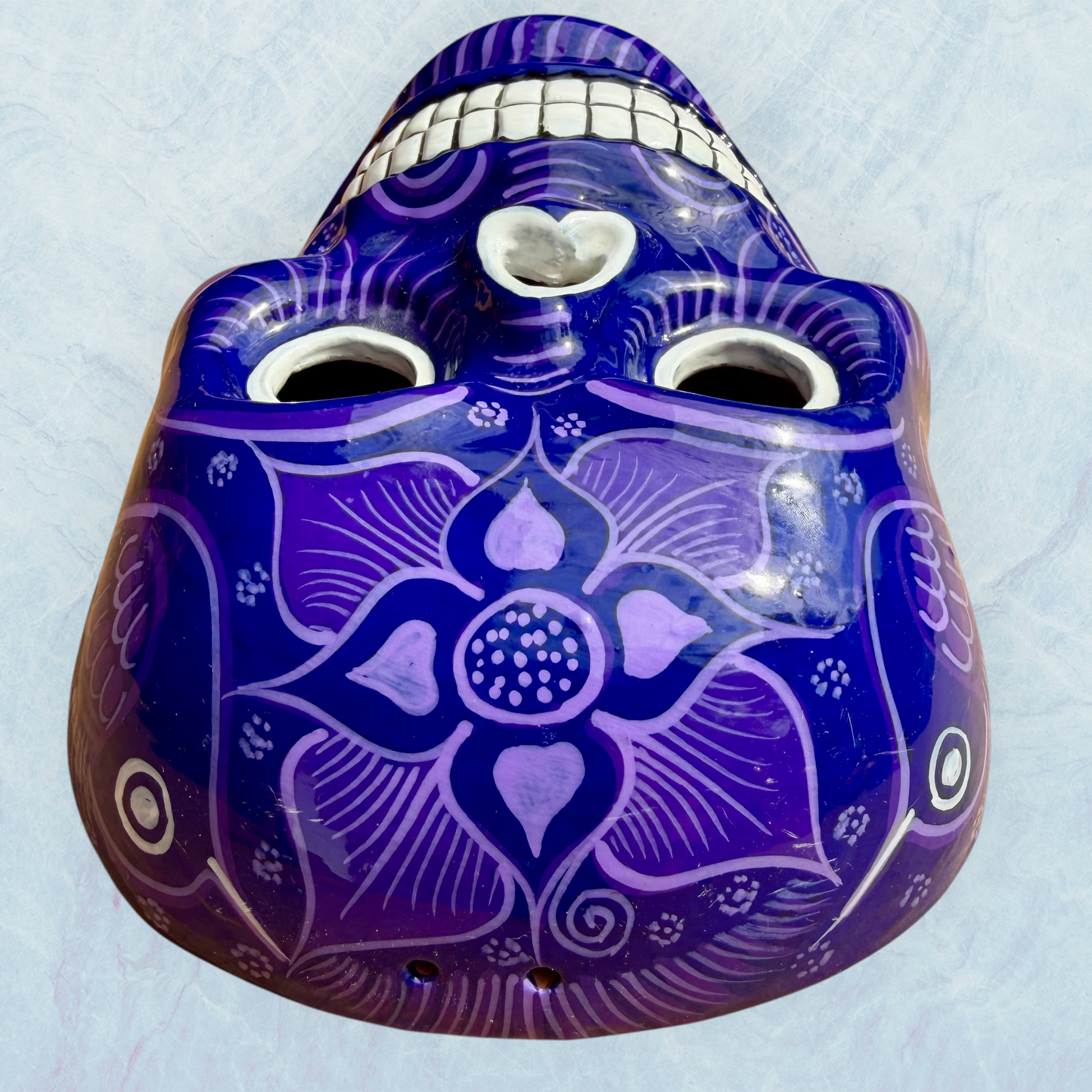 A clay, purple with designs, Mexican-style skeleton skull face mask hanging on a soft blue surface top forehead view.