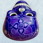 A clay, purple with designs, Mexican-style skeleton skull face mask hanging on a soft blue surface top forehead view.