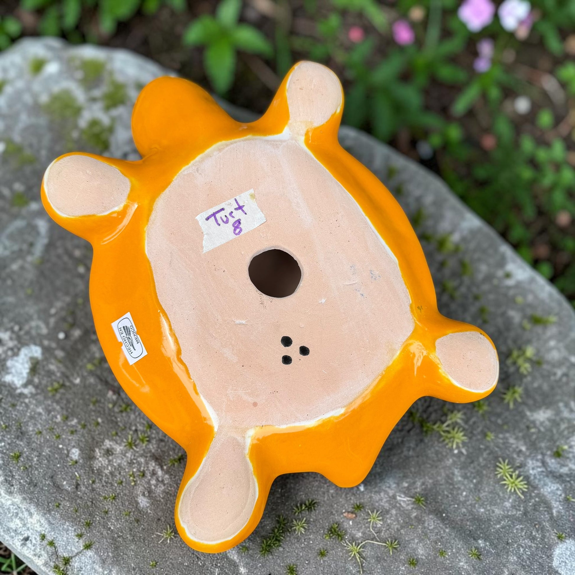 A glossy ceramic turtle figurine with a bright yellow body and a highly decorated shell in red, blue, yellow, black, and white, resting on a stone surface with a blurred garden background, bottom of turtle.