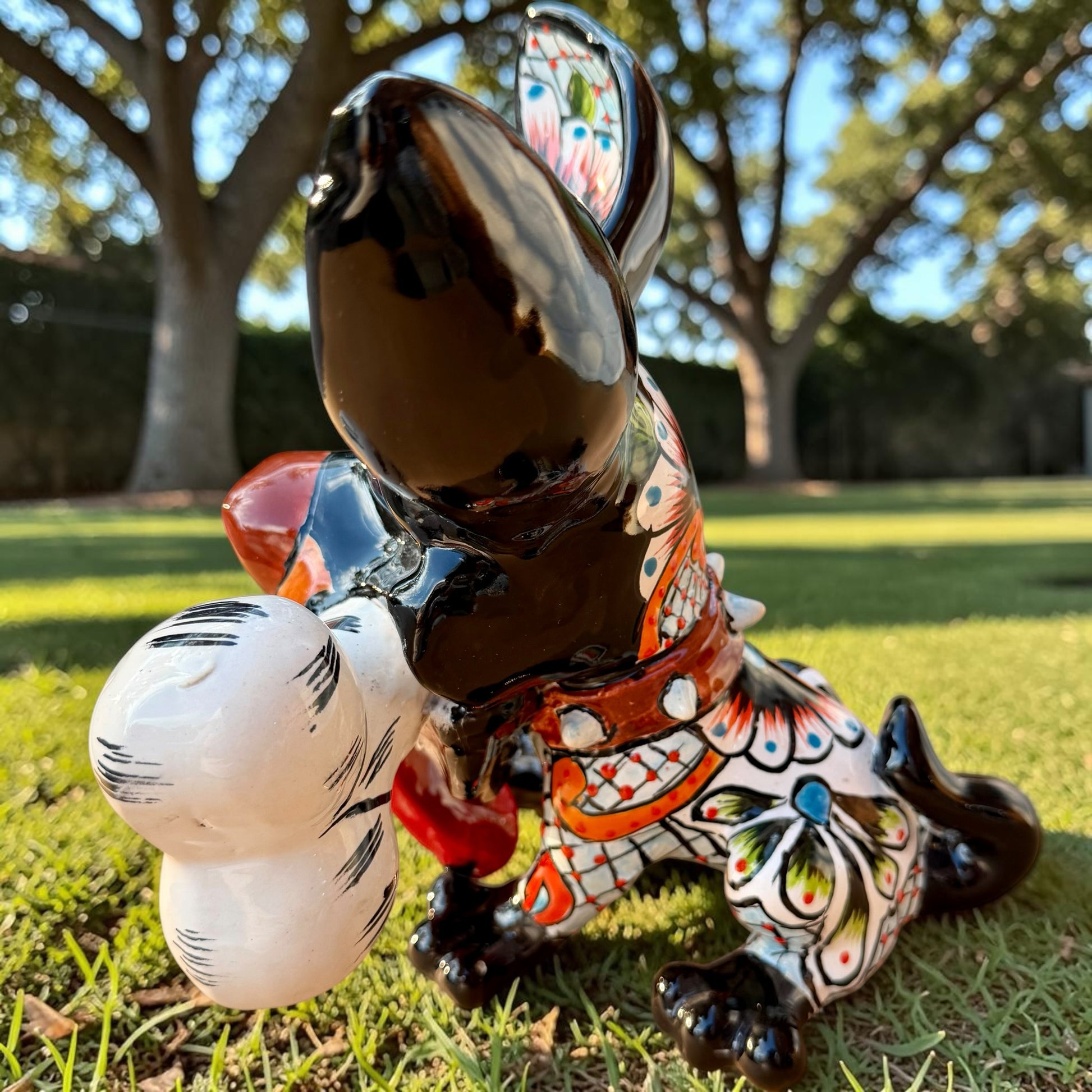 A colorful Talavera dog statue holding a big white bone, photographed outdoors on grass with trees in the background. The dog has a glossy black body, blue eyes, an orange snout, and multicolored patterned ears.  Right side view of the puppy dog.