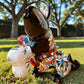 A colorful Talavera dog statue holding a big white bone, photographed outdoors on grass with trees in the background. The dog has a glossy black body, blue eyes, an orange snout, and multicolored patterned ears.  Right side view of the puppy dog.