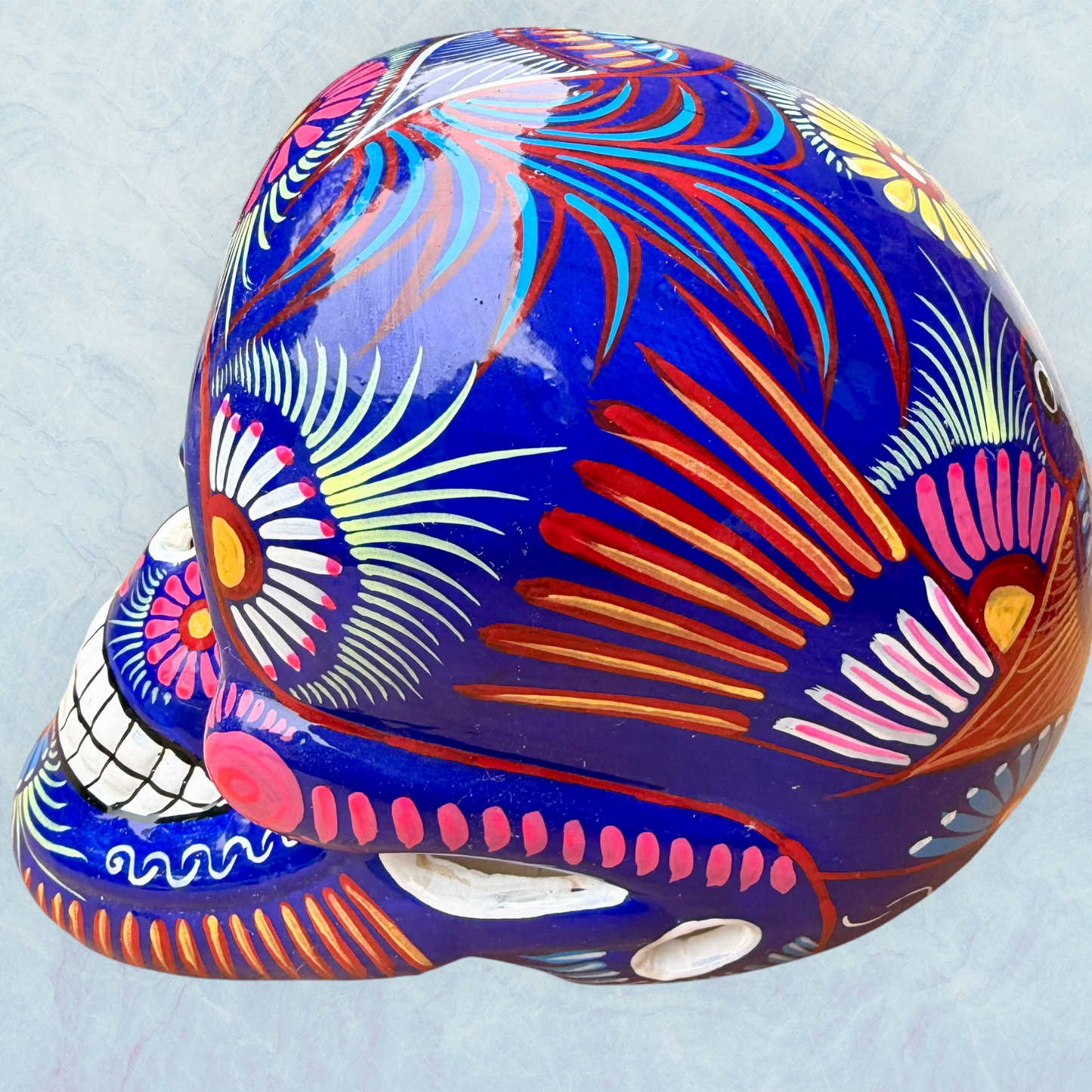 A glossy blue Day of the Dead sugar skull XGrande decorated with vibrant pink, blue, white, and orange floral and geometric patterns, shown against a light blue textured background, right side view.