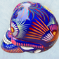 A glossy blue Day of the Dead sugar skull XGrande decorated with vibrant pink, blue, white, and orange floral and geometric patterns, shown against a light blue textured background, right side view.