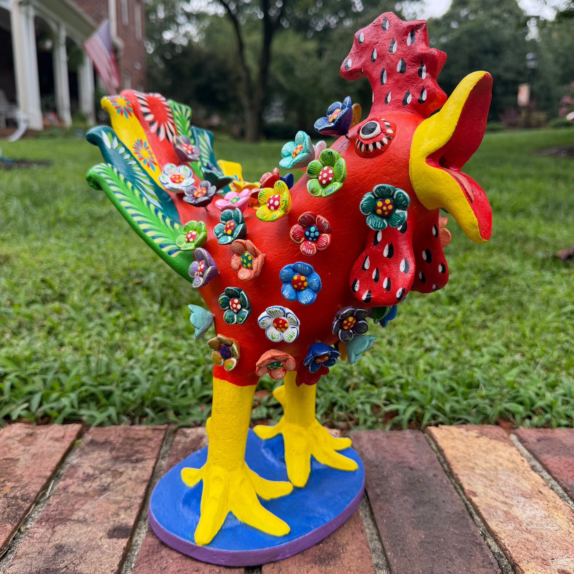 A colorful decorative rooster figurine standing left angle on a brick surface outdoors, with a grassy lawn and trees in the background. The rooster is primarily red and yellow with many small multicolored flowers and painted details across its body and tail.