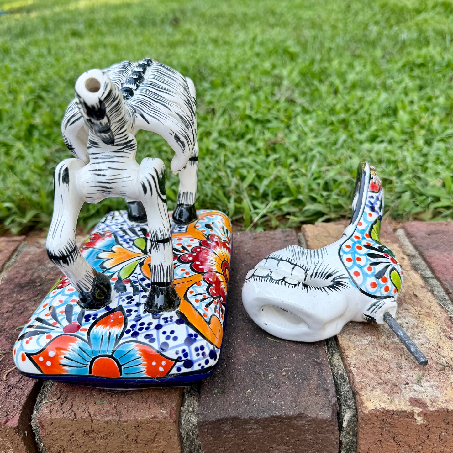 A hand-painted Mexican Talavera-style donkey figurine with a skeletal design, standing on a colorful floral base outdoors on a brick path with a green lawn in the background. This is the base with the head removed.