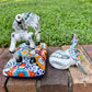 A hand-painted Mexican Talavera-style donkey figurine with a skeletal design, standing on a colorful floral base outdoors on a brick path with a green lawn in the background. This is the base with the head removed.