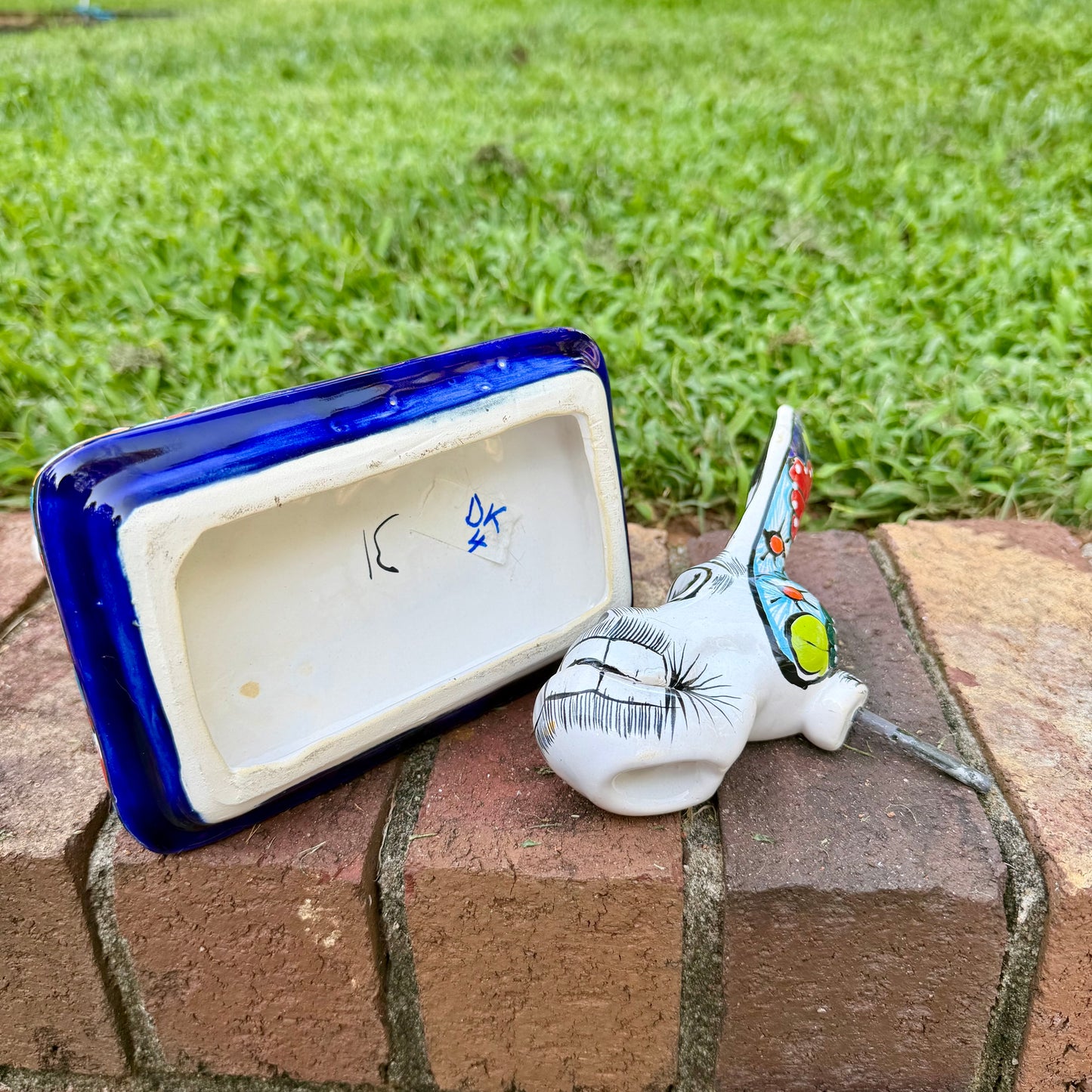 A colorful Catrina donkey figurine standing on a decorated base, photographed outdoors on a brick surface with a grassy yard and house in the background, bottem view. 
