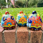 Three hand-painted Talavera ceramic jack-o'-lantern pumpkins with orange faces and yellow, blue, and red green designs, sitting on a brick path in a grassy backyard, back view.