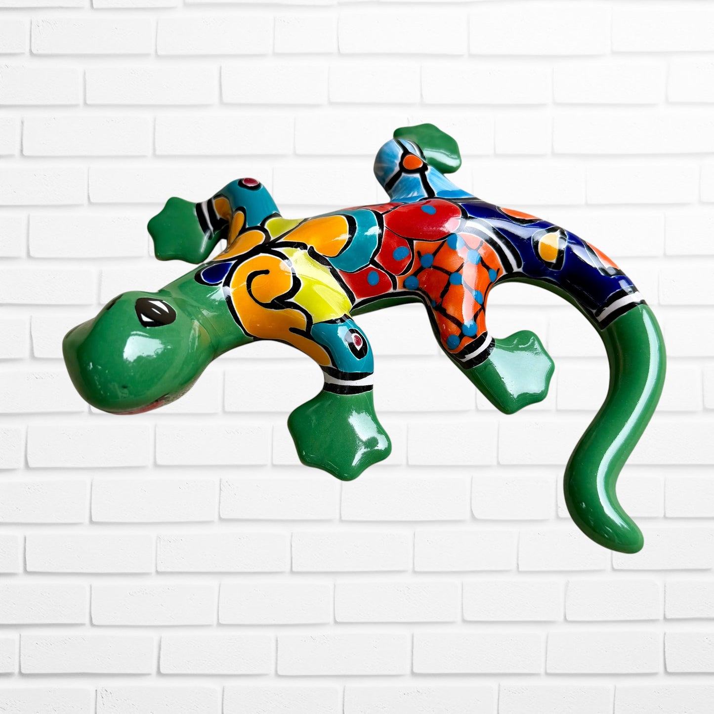 A colorful hand-painted ceramic lizard wall sculpture mounted on a white brick wall. The lizard features a vibrant mosaic pattern with green, blue, red, yellow, and orange sections front face leg view.