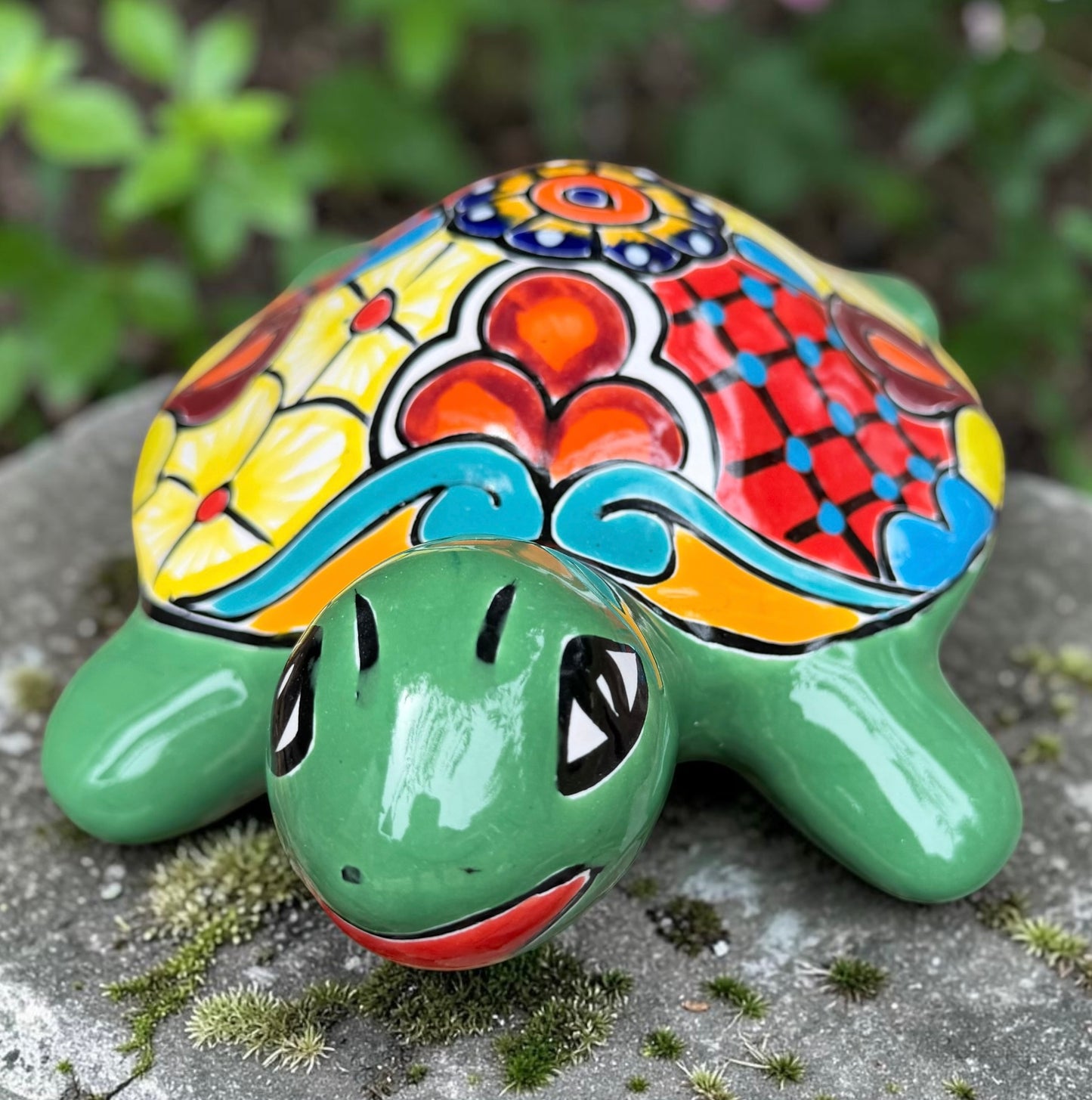 A hand-painted ceramic turtle figurine resting on a gray stone surface with a blurred green garden background. The turtle features a bright green head and legs, and a highly detailed, multicolored shell with yellow, red, orange, blue, and black patterns, front head view.
