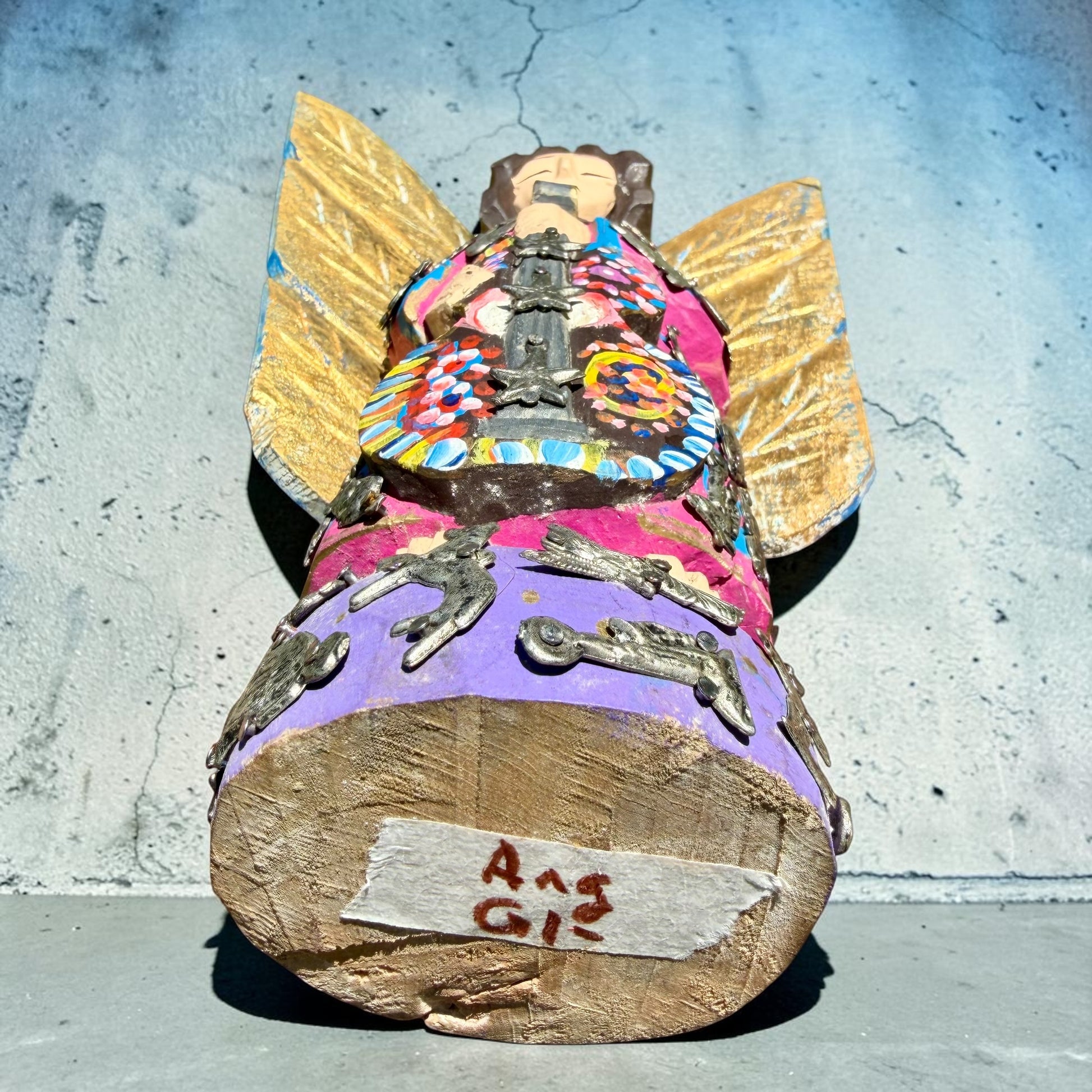 A handmade wood Milagros angel figurine with large gold wings, holding a guitar, dressed in a colorful patterned robe against a textured blue wall, bottom of sculpture.