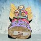 A handmade wood Milagros angel figurine with large gold wings, holding a guitar, dressed in a colorful patterned robe against a textured blue wall, bottom of sculpture.