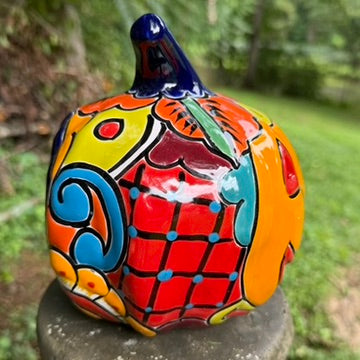 A hand-painted ceramic jack-o'-lantern pumpkin ornament with a bright orange face, red carved eyes and mouth, a blue stem, and colorful side patterns, sitting outdoors on a stone pedestal in a grassy yard left side view.
