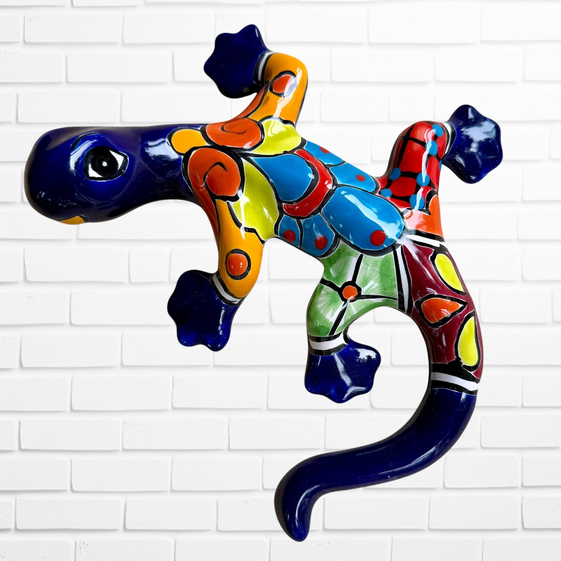 A colorful, glossy lizard wall sculpture with 
a vibrant mosaic pattern of blue, orange, yellow, green, red, and black, mounted on a white brick wall.