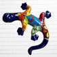 A colorful, glossy lizard wall sculpture with 
a vibrant mosaic pattern of blue, orange, yellow, green, red, and black, mounted on a white brick wall.