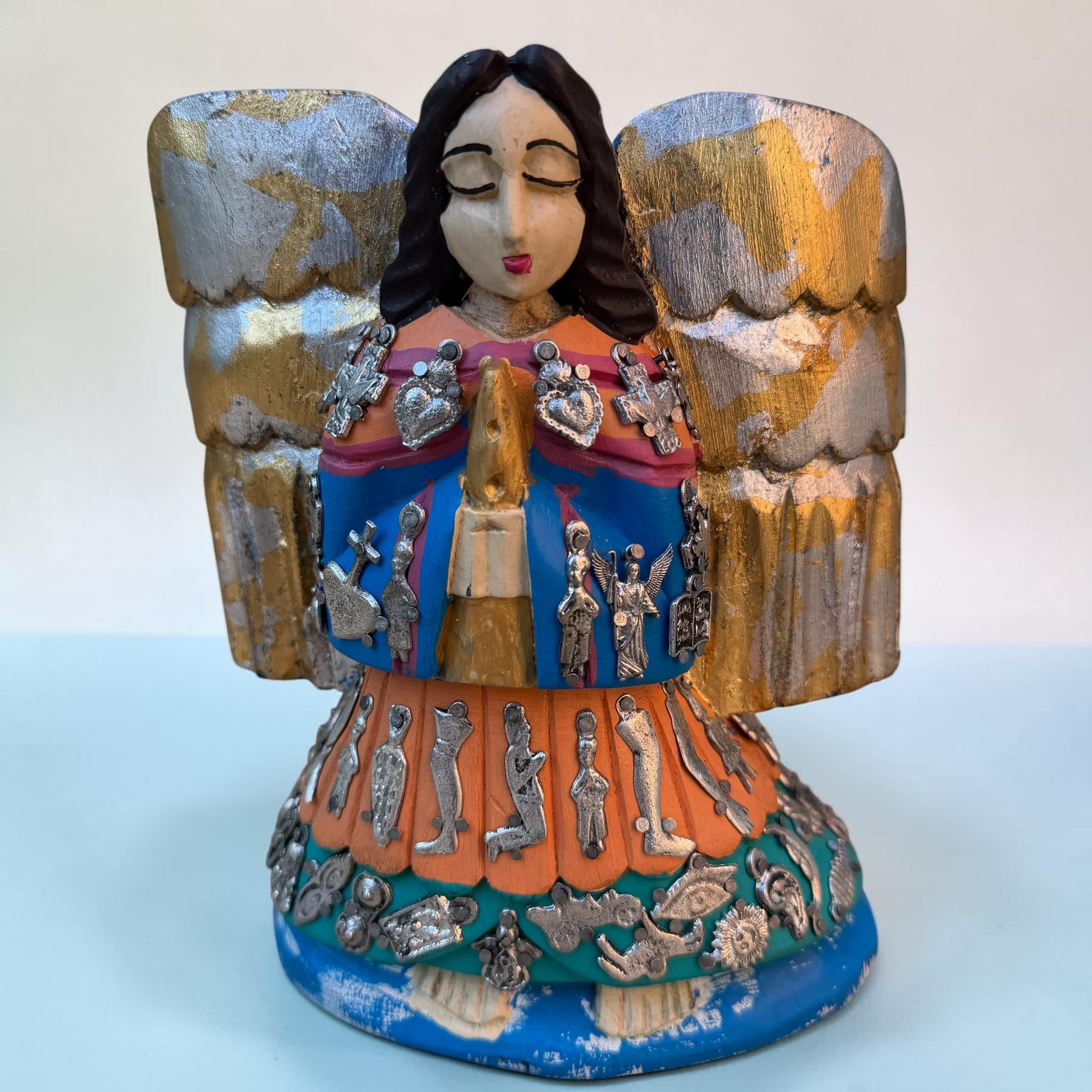 A colorful handcrafted Milagros angel figurine with large gold and silver wings, a blue and orange robe, and silver metallic embellishments, photographed against a plain light background.