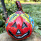 A hand-painted ceramic jack-o'-lantern pumpkin with a red face, black carved eyes and mouth, and a multicolored floral pattern on a garden background.