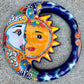 A colorful hand-painted talavera wall hanging shaped like a crescent moon and sun, featuring a glossy finish and vibrant orange, blue, yellow, and white details, displayed on a gray textured concrete surface.