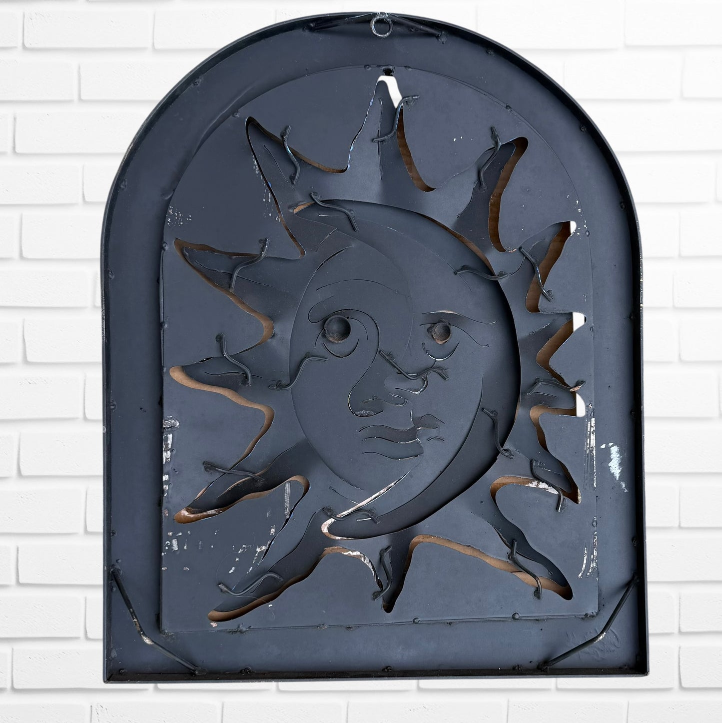 Decorative wall art of a sun and moon face with a metal frame on a white brick wall. Designed by artist Andres Martin back