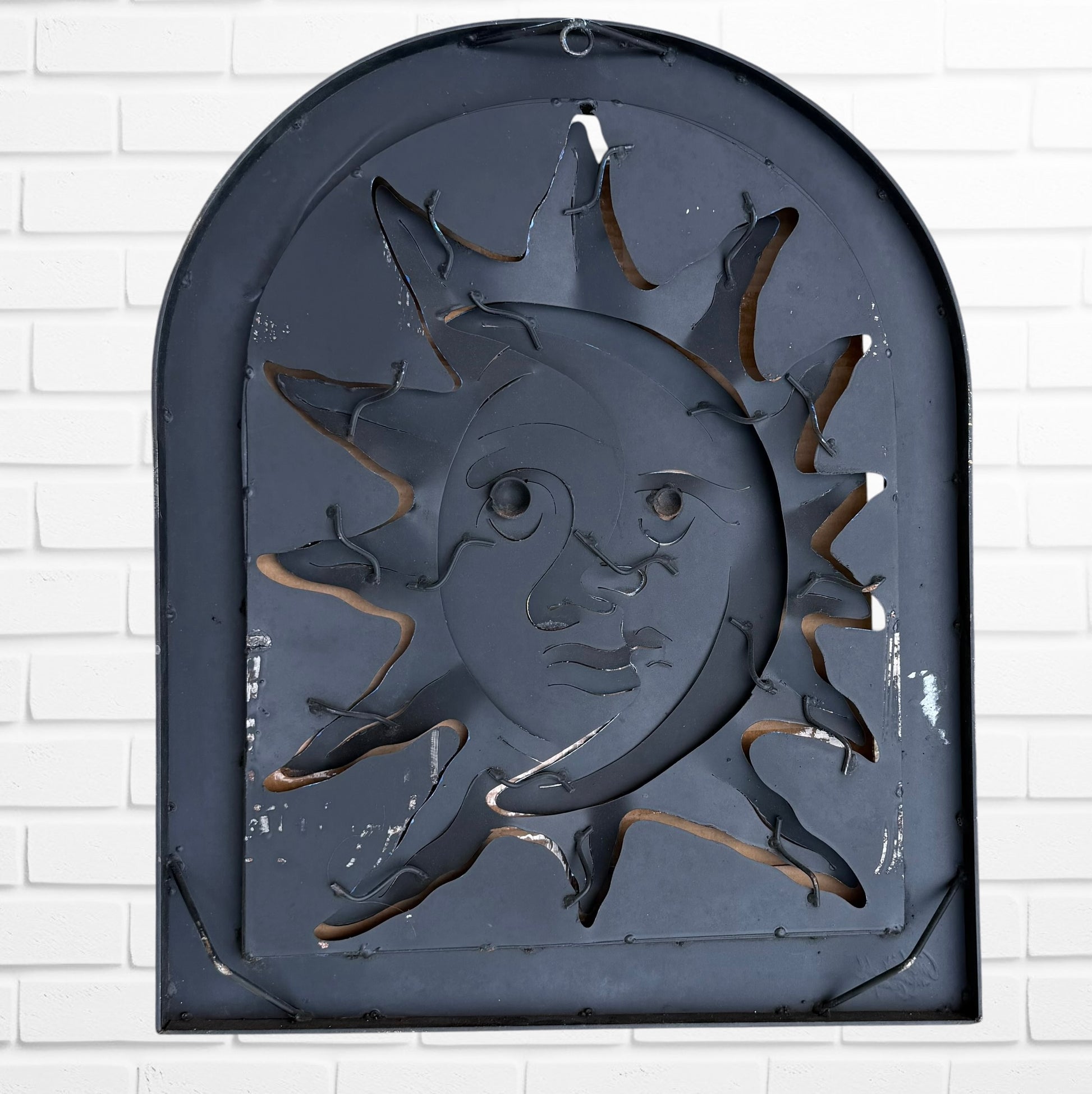 Decorative wall art of a sun and moon face with a metal frame on a white brick wall. Designed by artist Andres Martin back