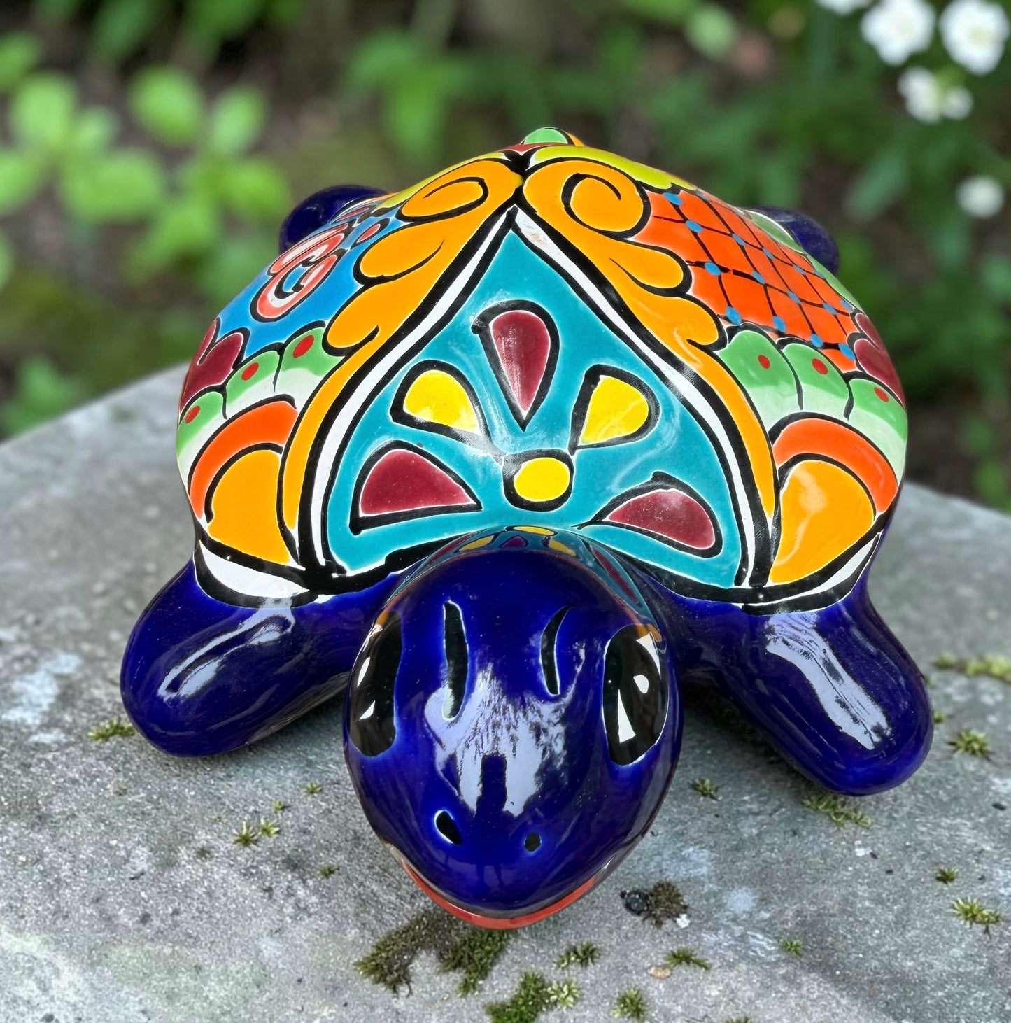 A glossy ceramic turtle figurine with a vibrant hand-painted patterned shell and a deep blue head and legs, resting on a gray stone surface with a blurred green garden background, front head view.