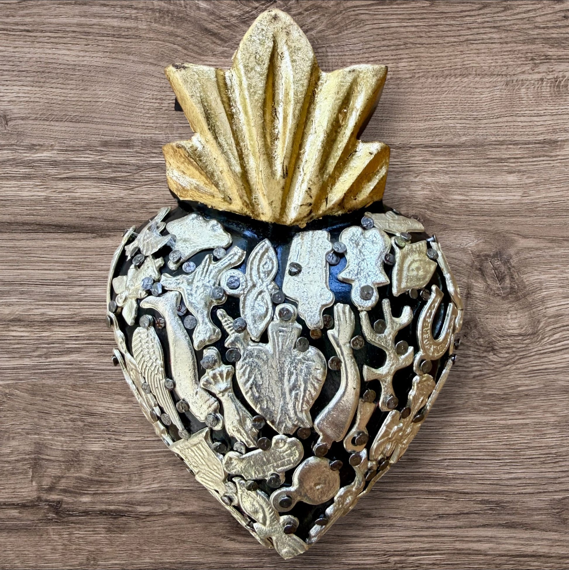 A Milagros heart featuring a gold textured top and a black heart base covered in raised silver-toned charms displayed on a wooden wall.