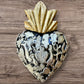 A Milagros heart featuring a gold textured top and a black heart base covered in raised silver-toned charms displayed on a wooden wall.