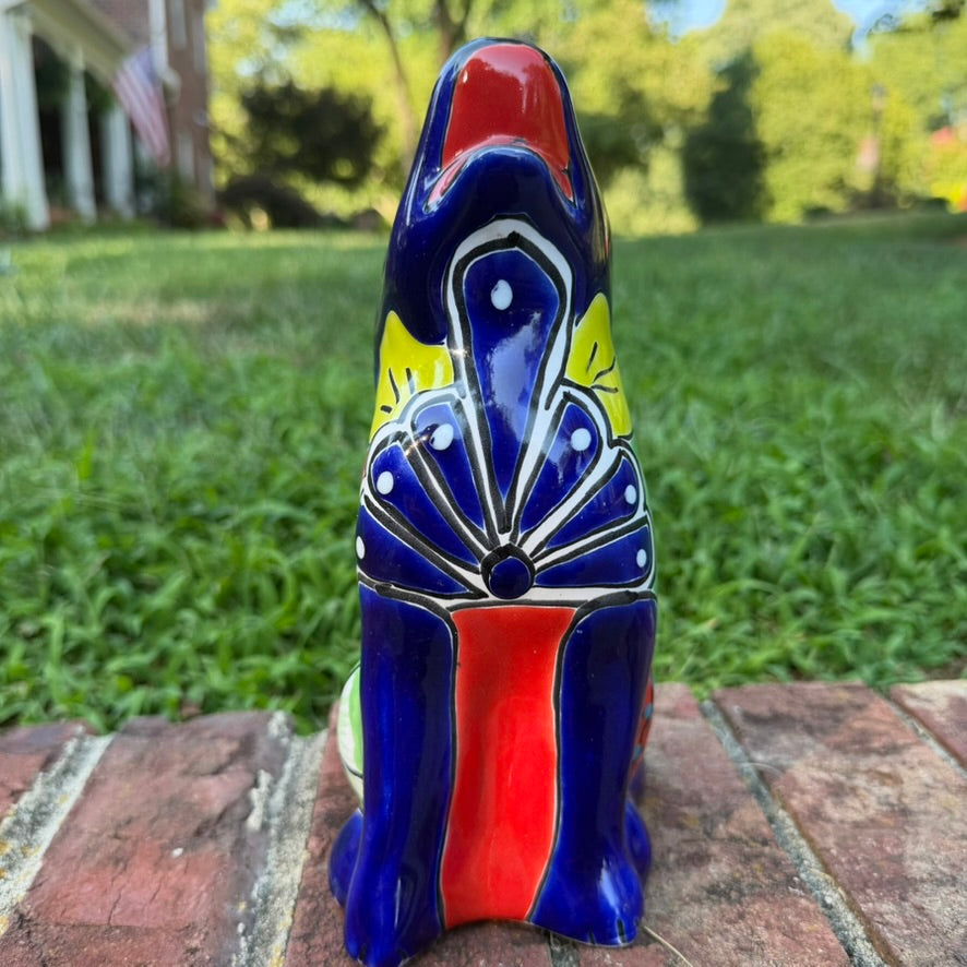 A colorful hand-painted talavera ceramic coyote figurine sitting on a brick surface with a blurred green lawn and trees in the background front view