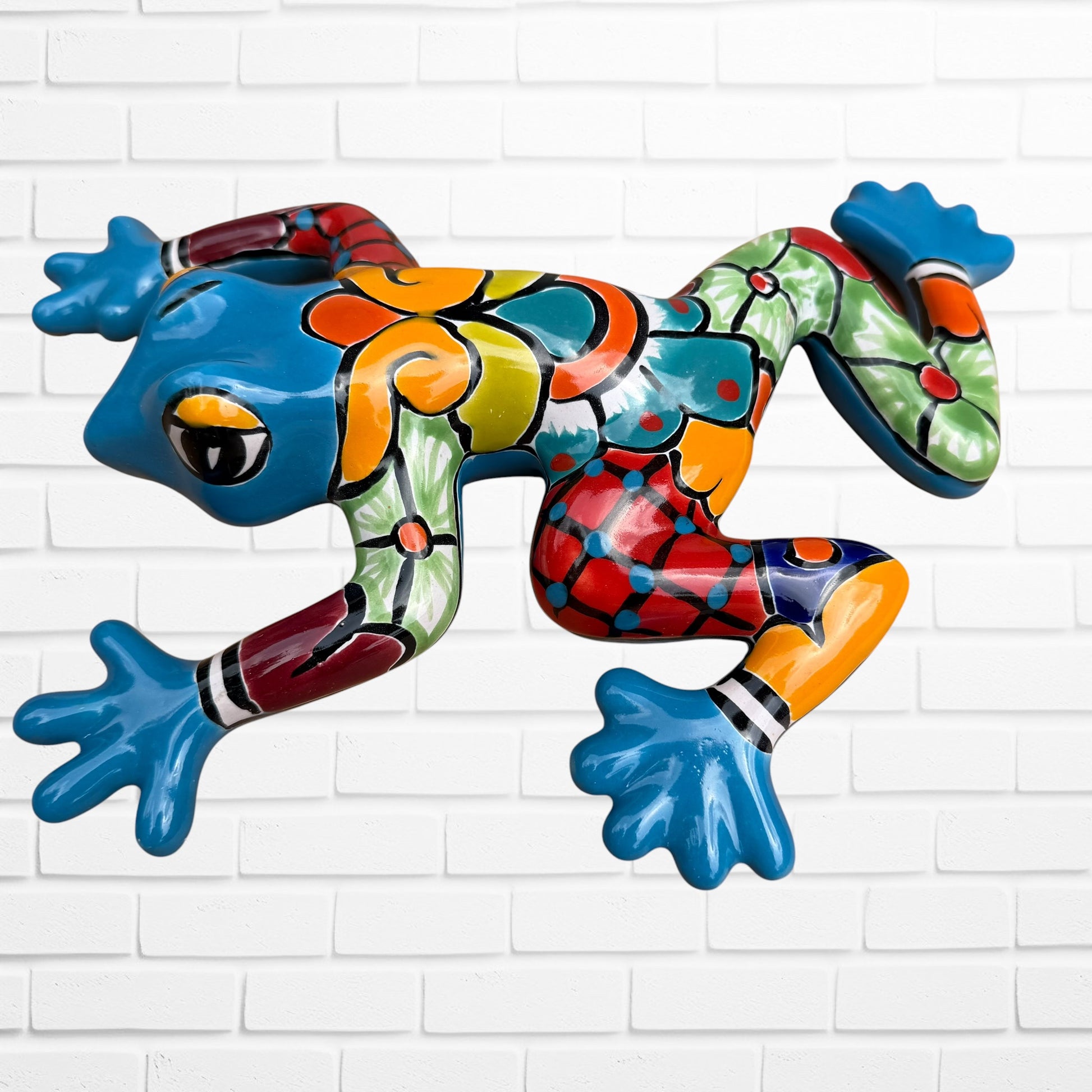 A colorful, glossy frog wall sculpture with a vibrant mosaic pattern in blue, green, orange, red, and yellow, mounted on a white brick wall.