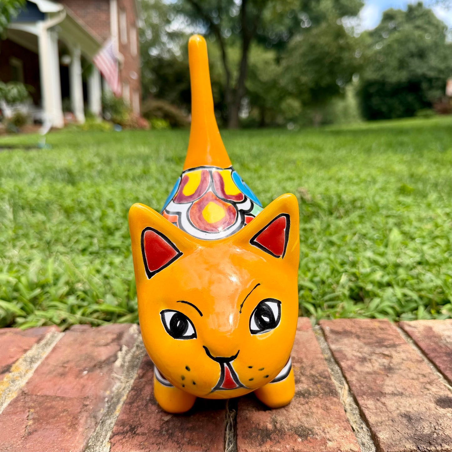 A hand-painted ceramic cat figurine in bright yellow with red, blue, and beige patterns, standing on a brick surface with a blurred green lawn and house in the background, Front face view.