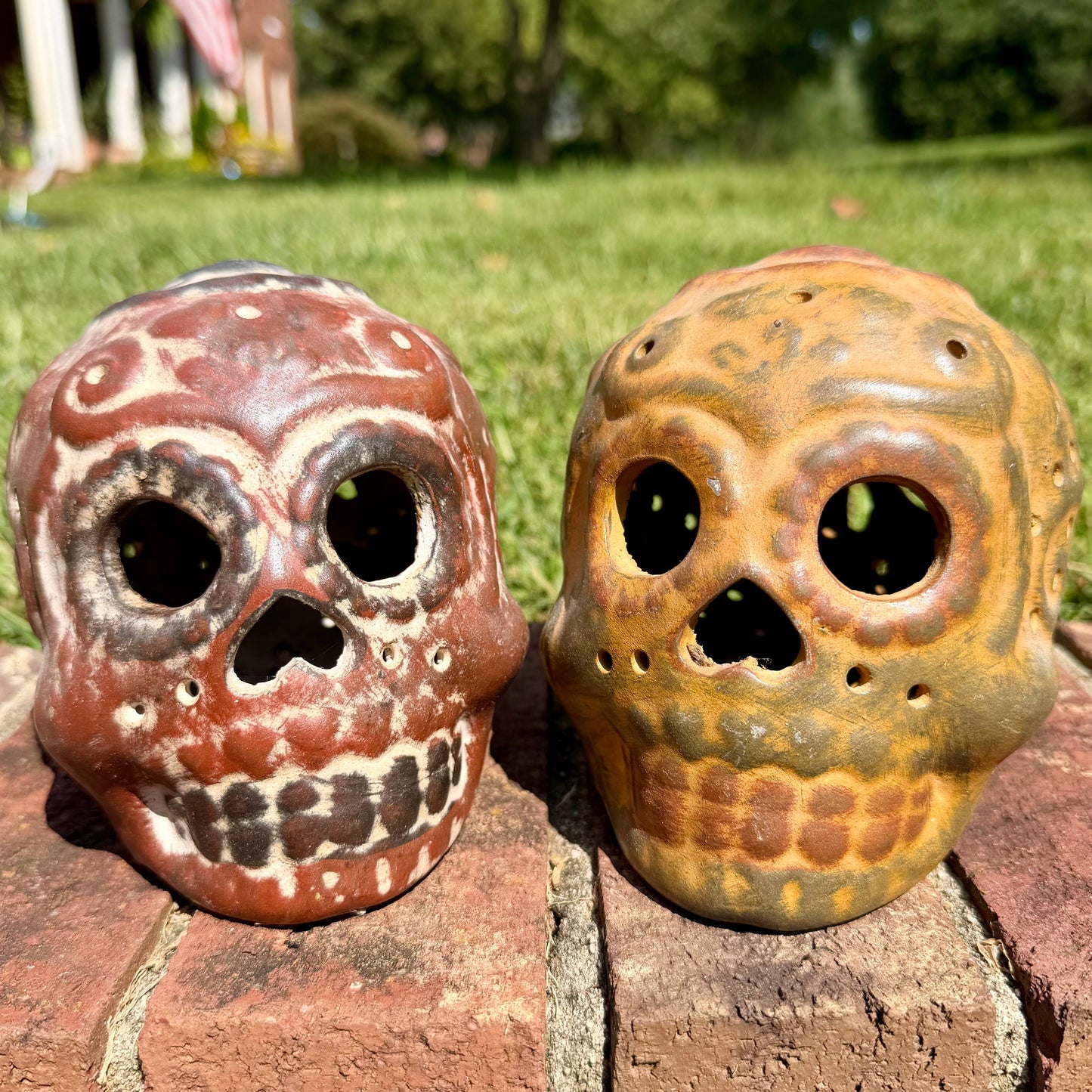 Two decorative clay sugar skull ornaments with Day of the Dead style painted patterns, displayed side-by-side on a brick surface outdoors with a grassy lawn and trees in the background.