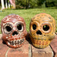 Two decorative clay sugar skull ornaments with Day of the Dead style painted patterns, displayed side-by-side on a brick surface outdoors with a grassy lawn and trees in the background.