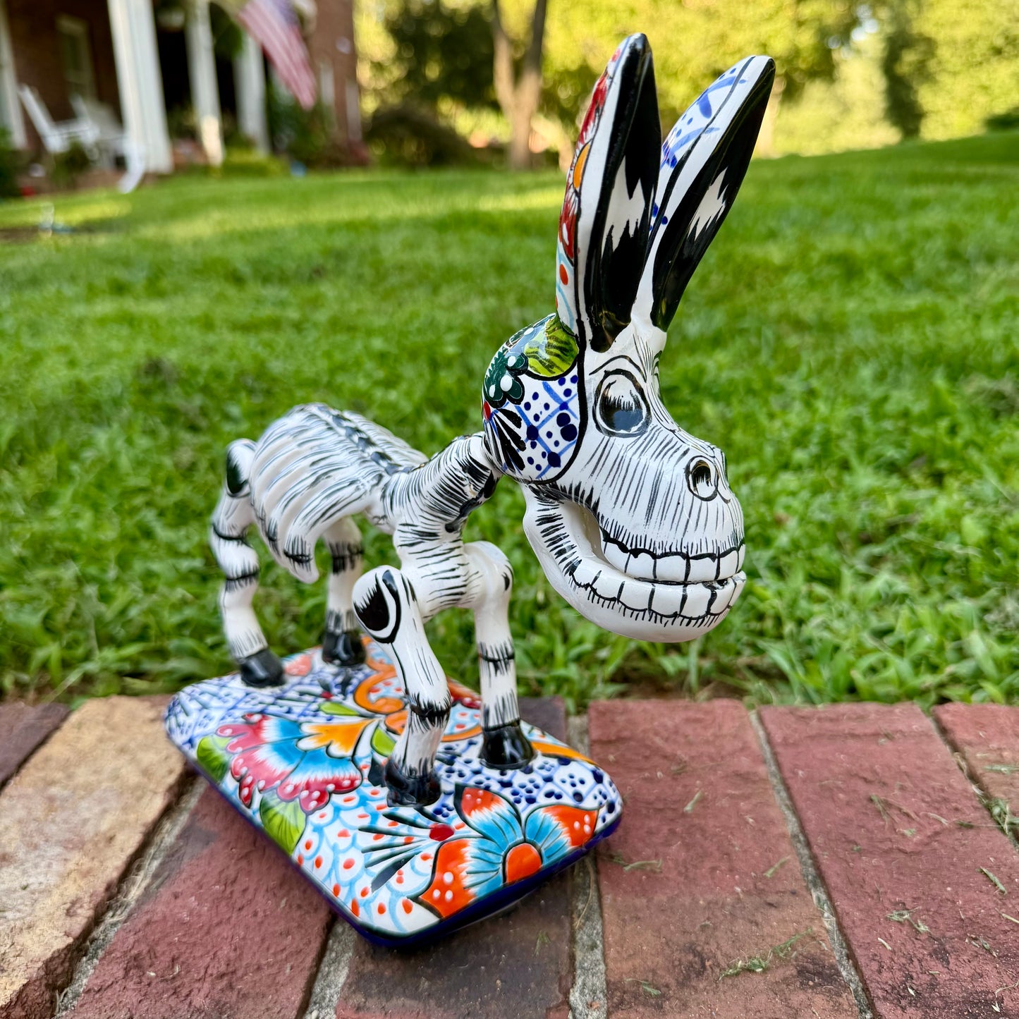A hand-painted Mexican Talavera-style donkey figurine with a skeletal design, standing on a colorful floral base outdoors on a brick path with a green lawn in the background.