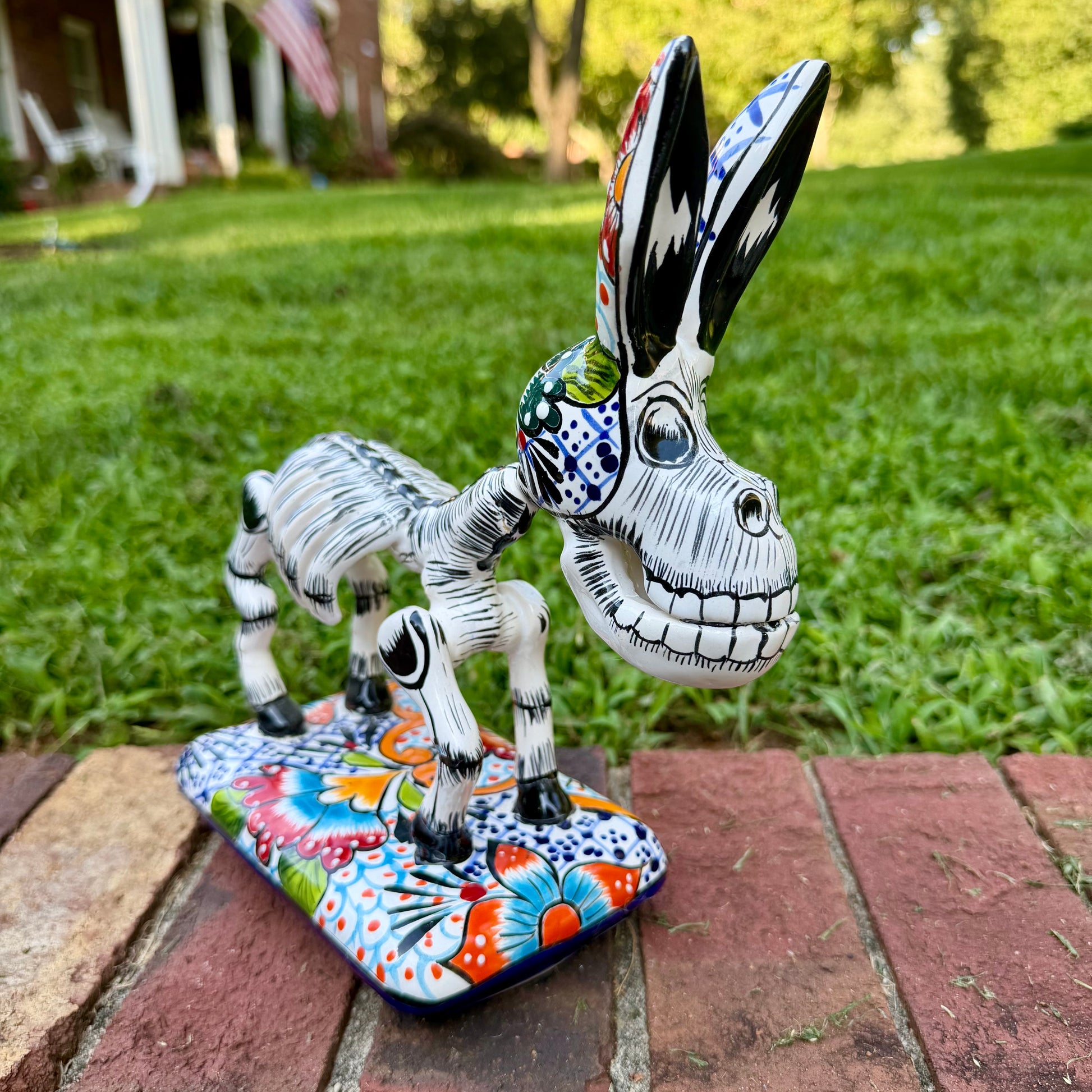 A hand-painted Mexican Talavera-style donkey figurine with a skeletal design, standing on a colorful floral base outdoors on a brick path with a green lawn in the background.