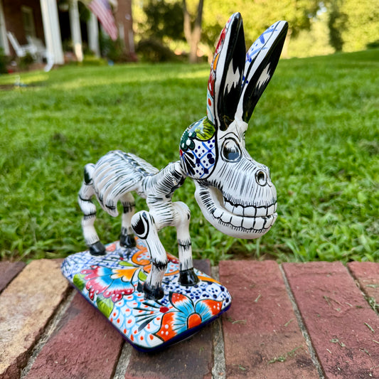 A hand-painted Mexican Talavera-style donkey figurine with a skeletal design, standing on a colorful floral base outdoors on a brick path with a green lawn in the background.