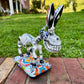 A hand-painted Mexican Talavera-style donkey figurine with a skeletal design, standing on a colorful floral base outdoors on a brick path with a green lawn in the background.