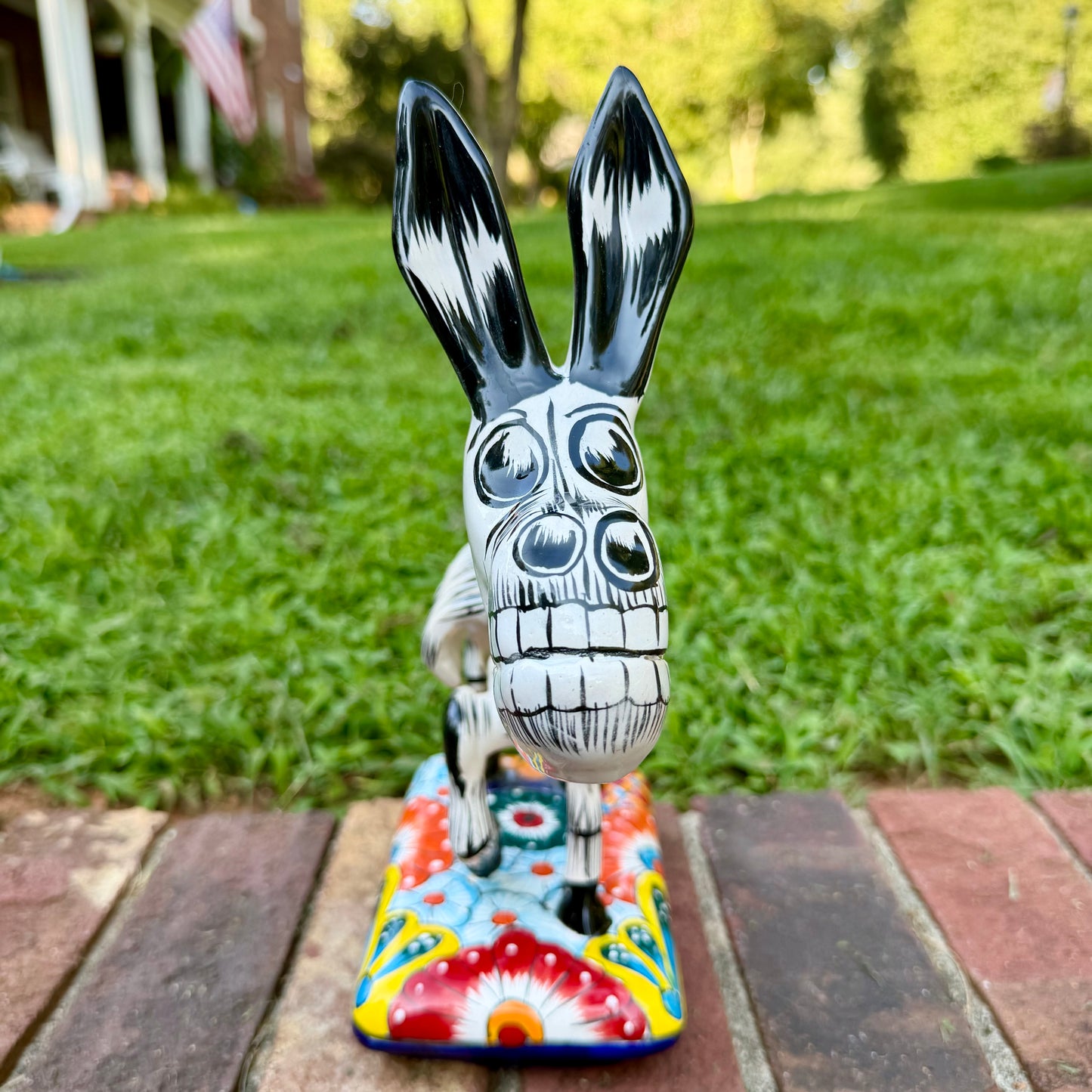 A colorful hand-painted Talavera Catrina donkey figurine, standing on a colorful floral base in an outdoor backyard setting, Front view showing face.
