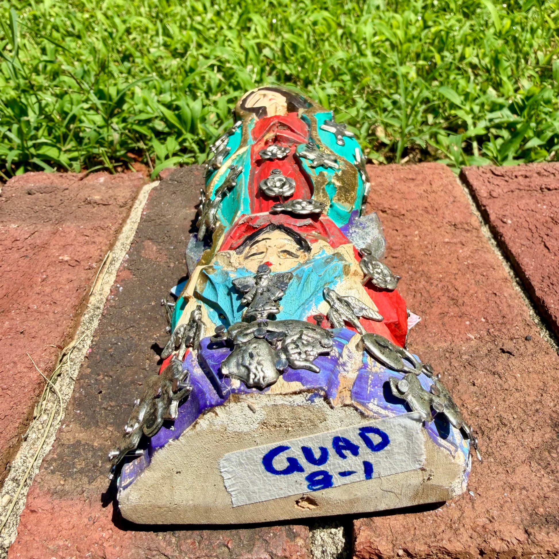 A colorful Milagros Guadalupe  (Virgin Mary) standing on a brick surface outdoors with a grassy yard in the background, bottom view.