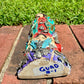 A colorful Milagros Guadalupe  (Virgin Mary) standing on a brick surface outdoors with a grassy yard in the background, bottom view.