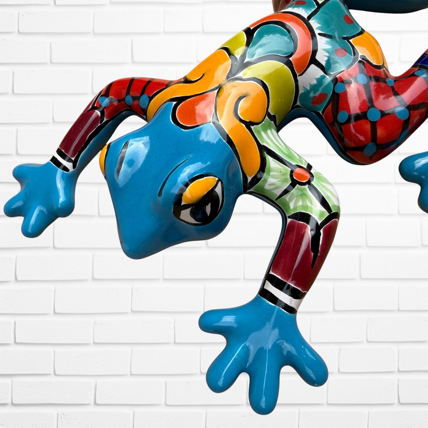 A colorful painted frog sculpture shown against a white brick wall background. The frog features a glossy blue head and hands, with red, orange, yellow, green, and black patterned details across its body.