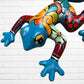 A colorful painted frog sculpture shown against a white brick wall background. The frog features a glossy blue head and hands, with red, orange, yellow, green, and black patterned details across its body.