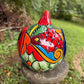 A hand-painted talavera Halloween  jack-o'-lantern candle holder in red, green, black, and orange, displayed on a stone pedestal in an outdoor garden setting. This is the right side view.