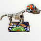 A ceramic Mexican-style skeleton dog figurine standing on a colorful floral base against a plain white background.  Left side view of catrina skeleton dog.