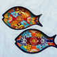Two decorative fish-shaped talavera serving plates with colorful hand-painted floral and geometric patterns, displayed on a light blue textured background.