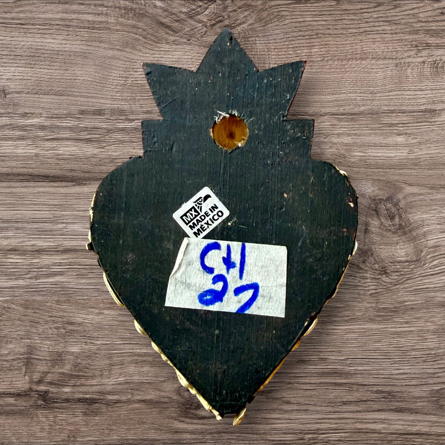 A decorative Milagros Sacred Heart featuring a black heart, red flame and antiqued charms, set against a rustic wood background. This is the back view.