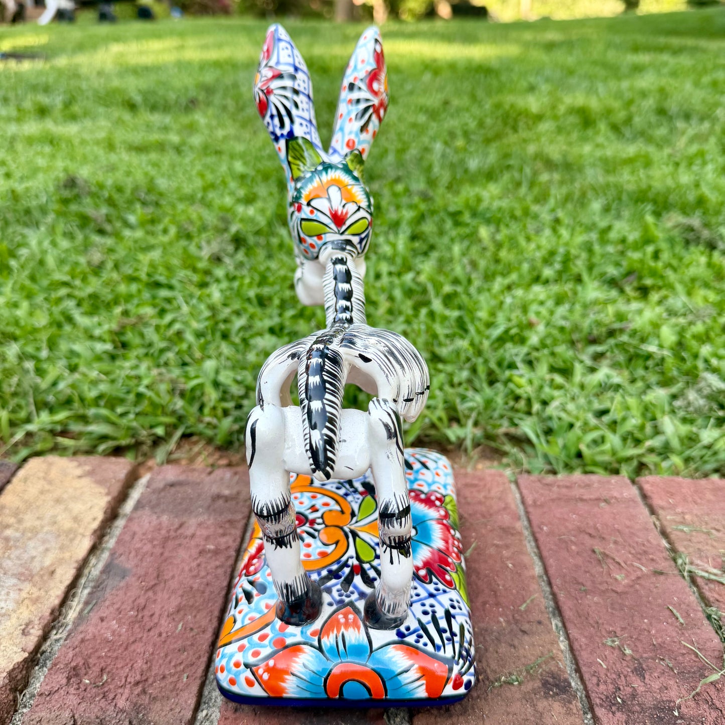 A hand-painted Mexican Talavera-style donkey figurine with a skeletal design, standing on a colorful floral base outdoors on a brick path with a green lawn in the background. Back view of Donkey.