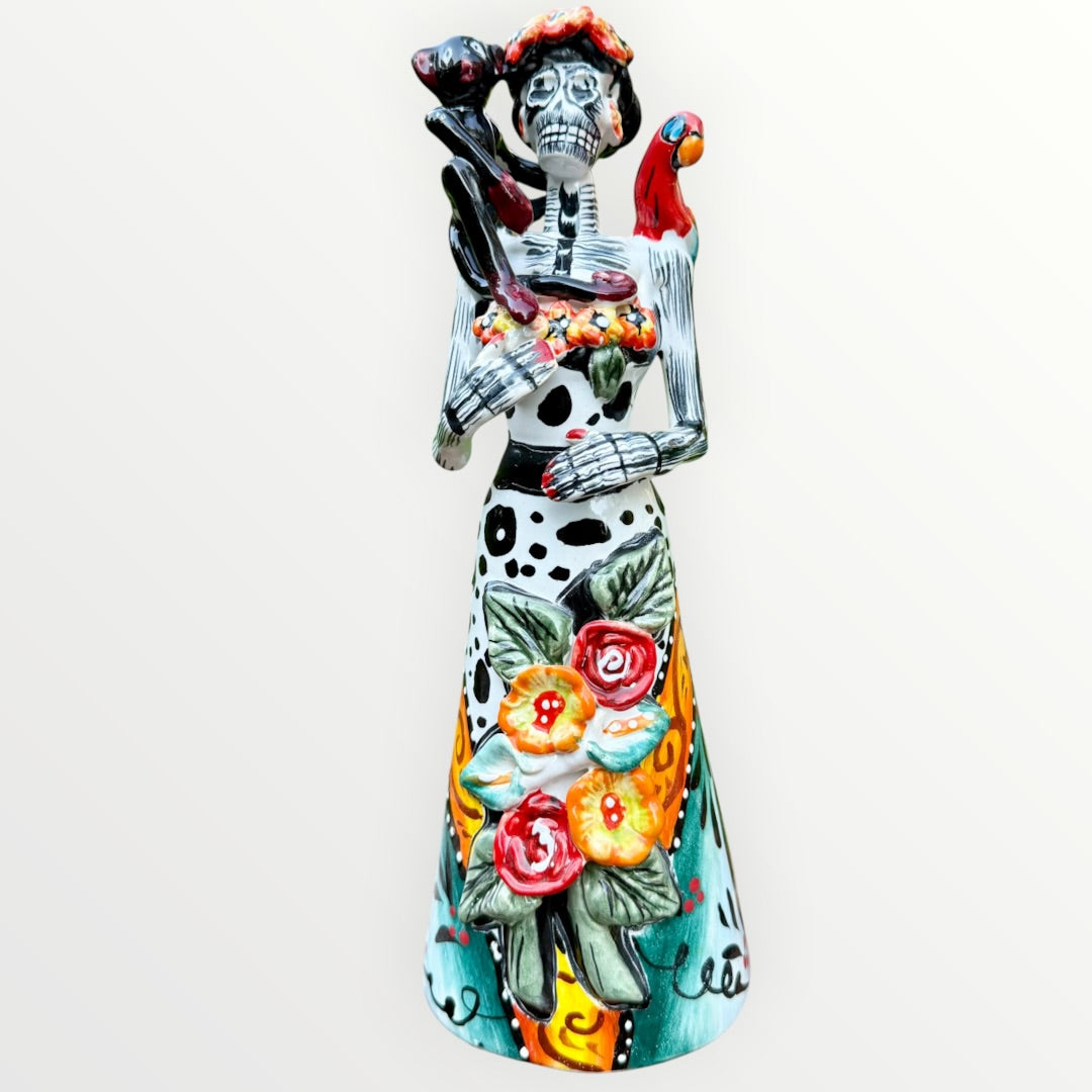 A Day of the Dead Catrina of a Frida skeleton doll holding a black cat, with a red parrot on her shoulder and colorful flowers around the top of her dress. Front view of doll.