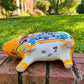 A colorful ceramic pig planter with a yellow-orange face and body, decorated with blue, green, red, and white floral patterns, sitting on a brick surface with a blurred outdoor garden background. Bottom of pig showing drain hole. 